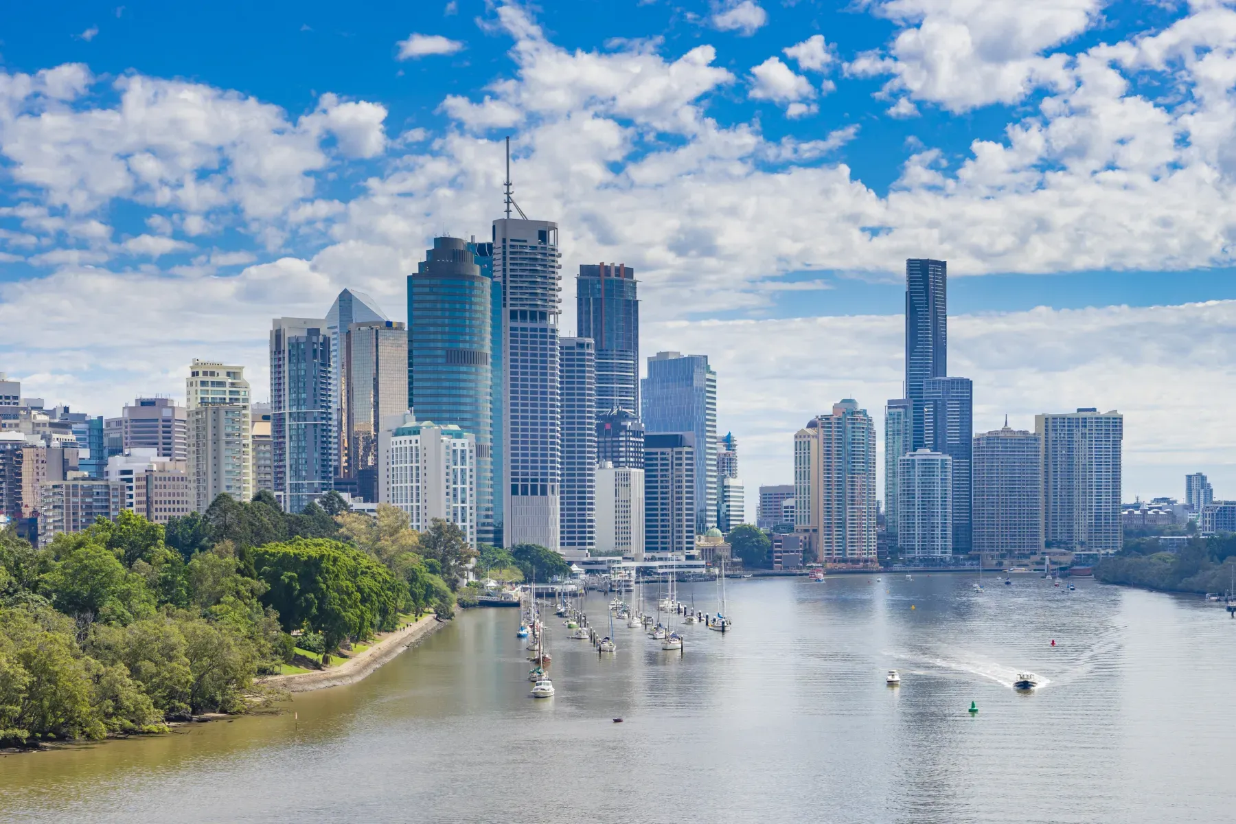 Brisbane city skyline along the Brisbane River, with boats moored and cruising on the water under a blue sky